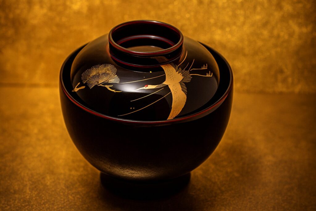 Black lacquer Japanese bowl with gold-painted crane and pine tree design, displayed against a radiant golden background