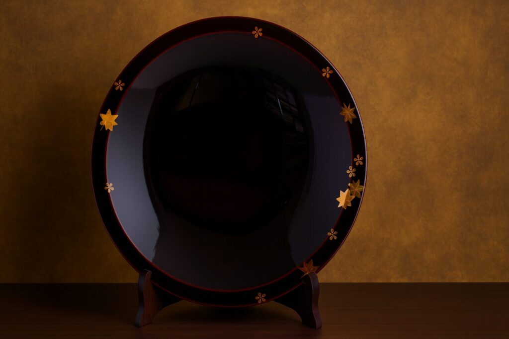 Round black lacquer plate with gold maple leaf and cherry blossom motifs, displayed on a wooden stand against a golden background