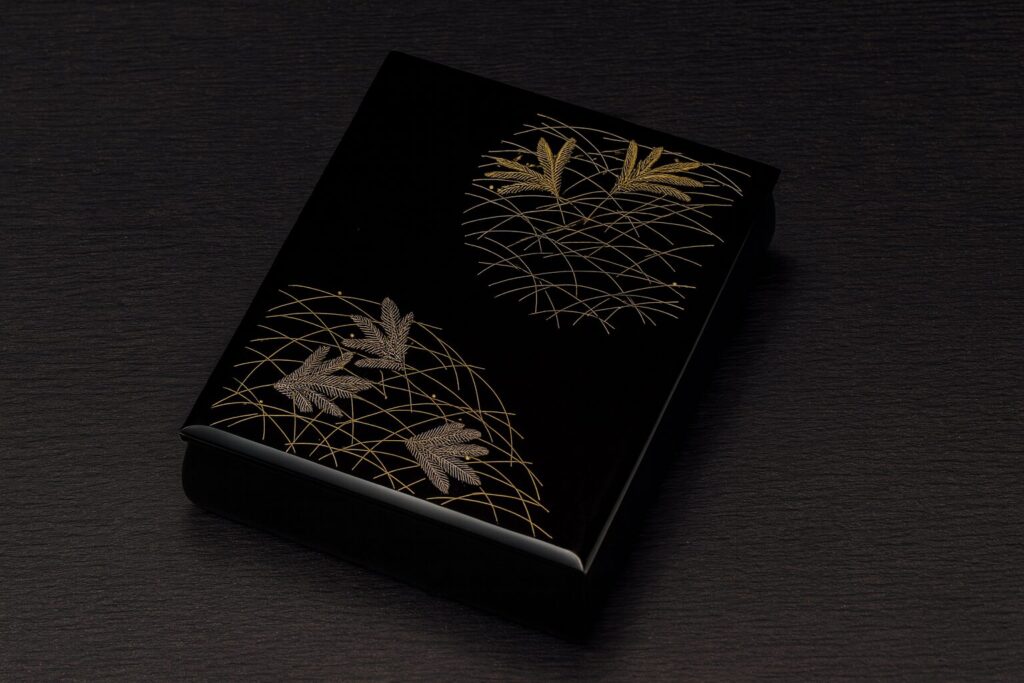 Black lacquer box with intricate gold pine needle and bamboo leaf design on a dark wood surface