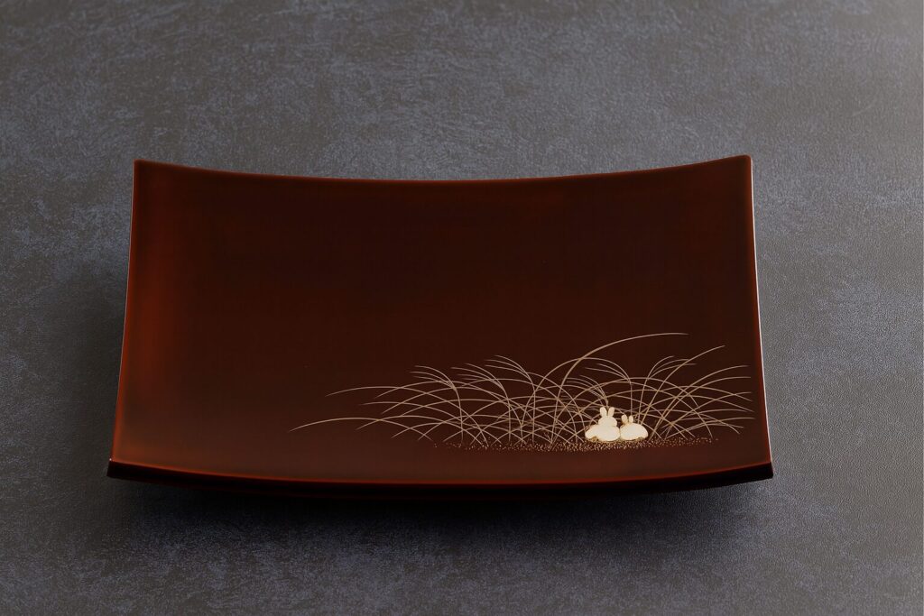 Rectangular brown lacquer plate with gold rabbit and grass motif, displayed on a dark textured surface