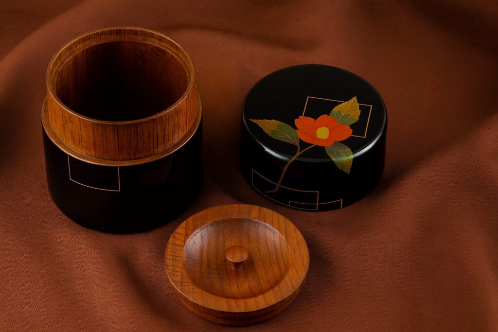 Black lacquer tea caddy with red camellia design and wooden interior on a brown fabric background