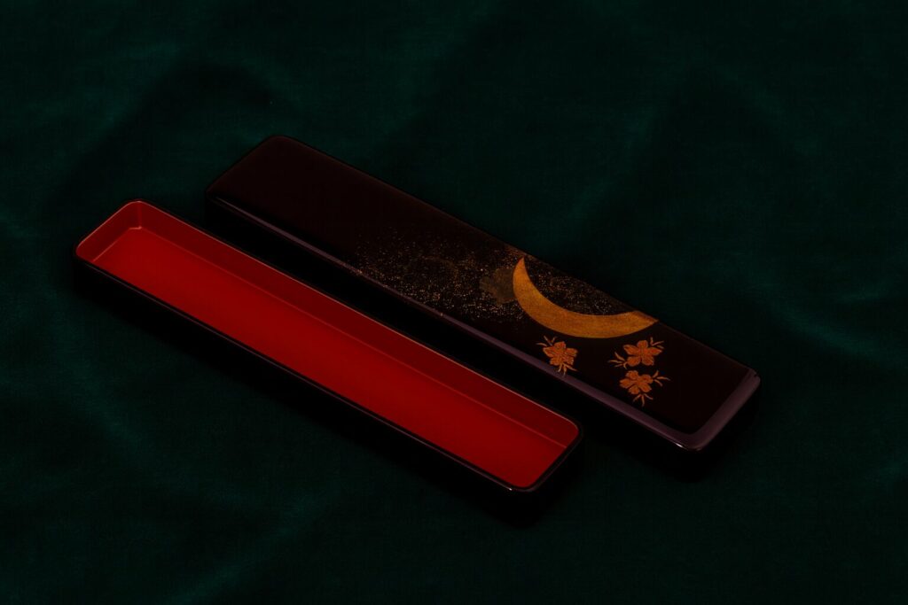 Black lacquer chopstick case with gold crescent moon and cherry blossom design, red interior, on a dark green fabric background